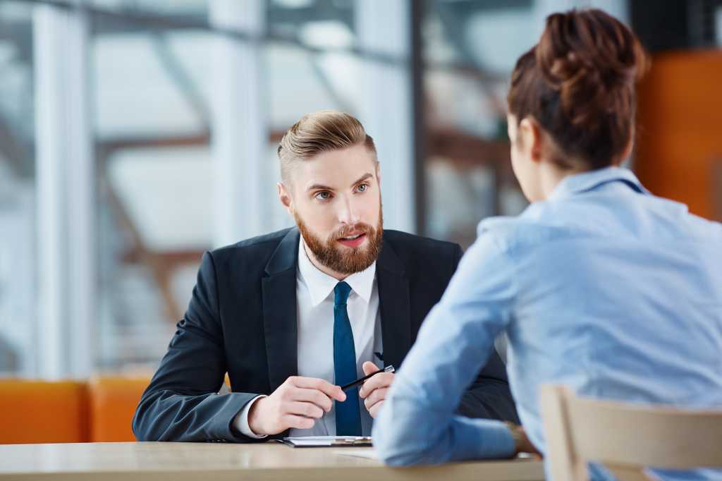 A man and woman sit on opposite sides of an office desk, in discussion.